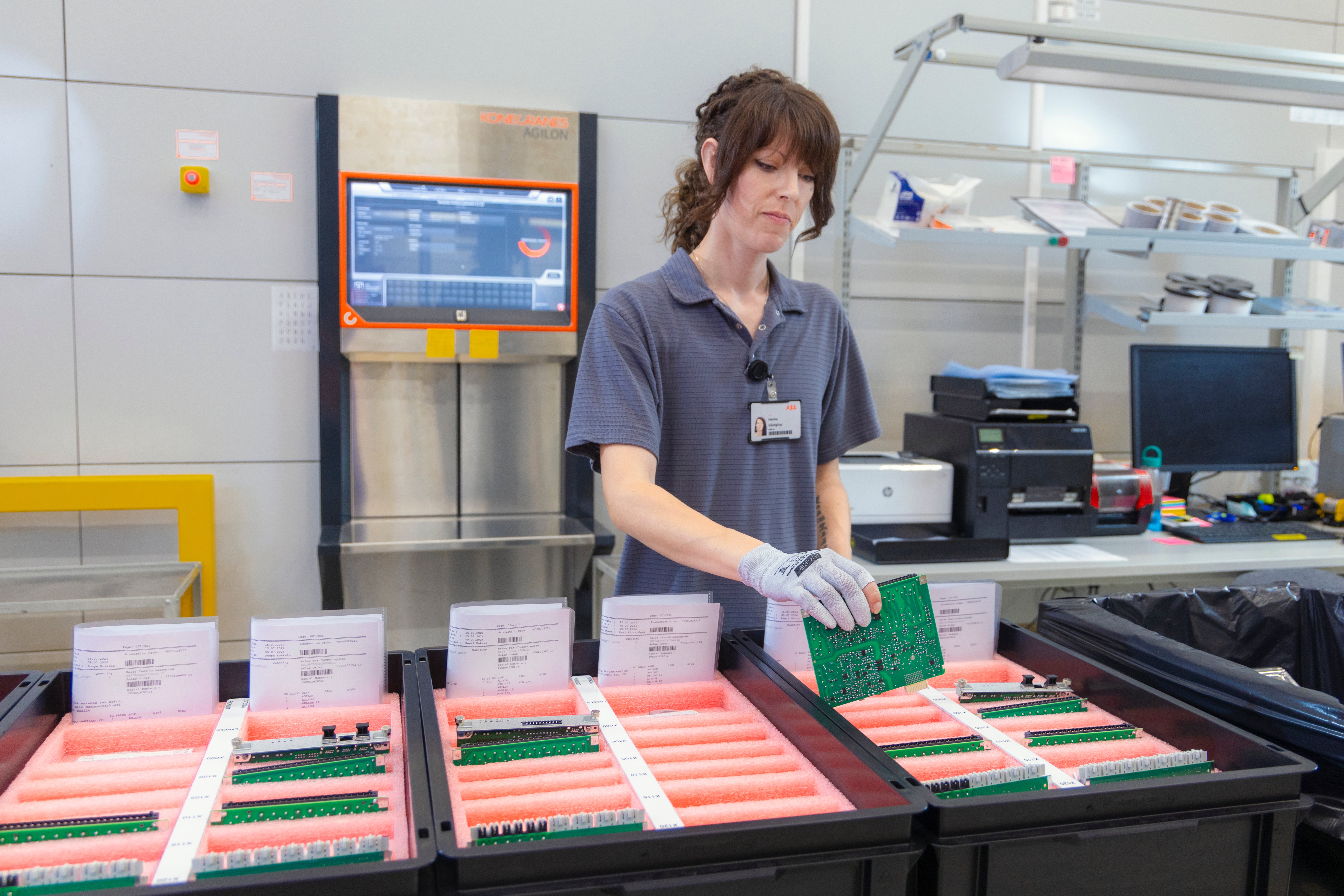 ABB Distribution Solutions' production picking process with Konecranes Agilon WMS. The image shows a woman picking parts for relay assembly. Behind her is the Agilon warehouse unit and user access point, while in front is an ergonomic PTL (Pick-to-Light) table with picking boxes containing already picked components and work order prints.